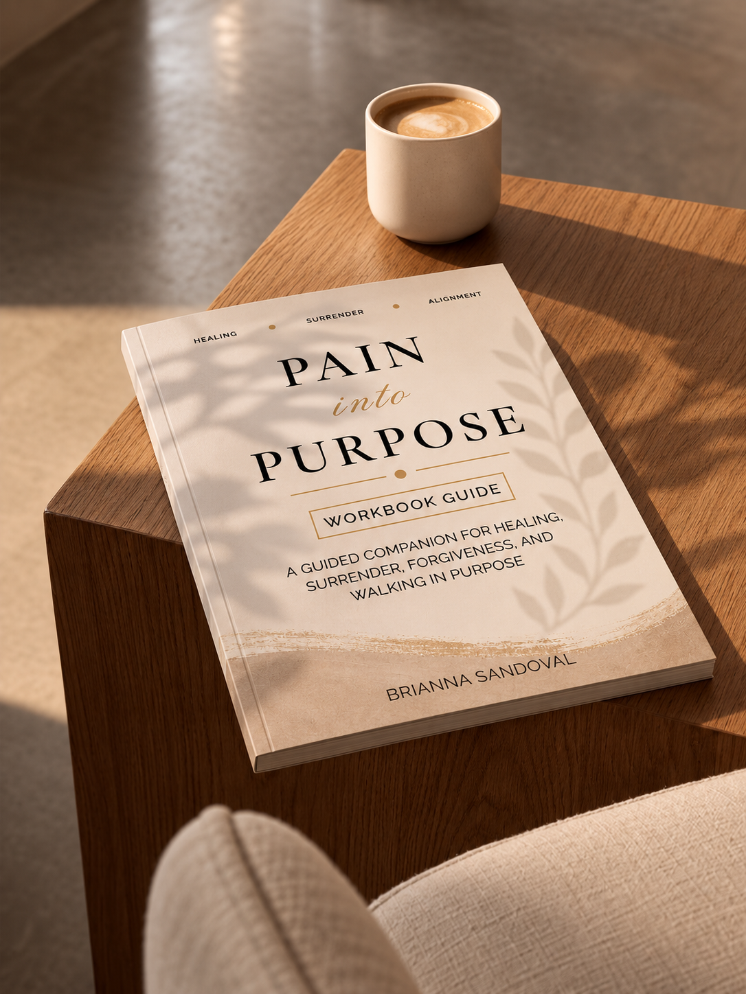 A book titled "Pain into Purpose: Workbook Guide" by Brianna Sandoval lies on a wooden table next to a cup of coffee, with soft leaf shadows cast across the cover.