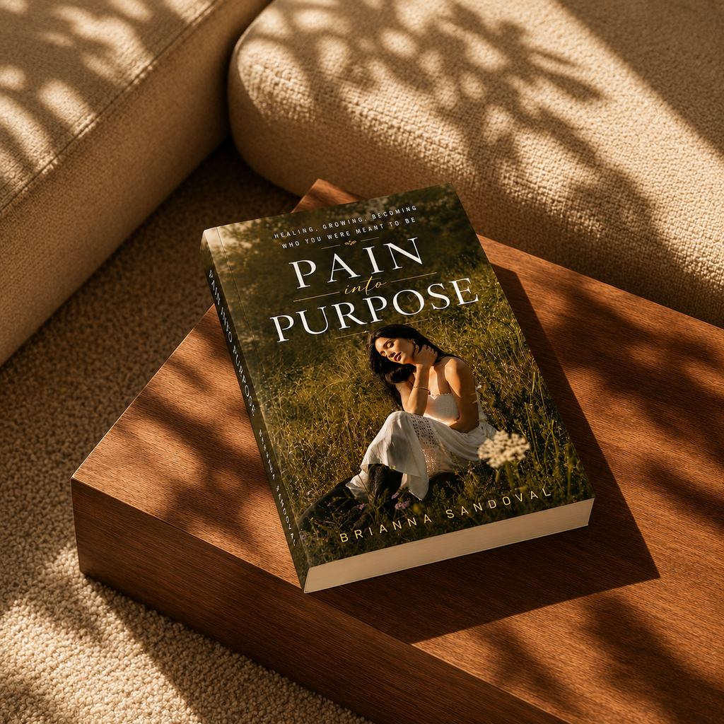 A hardcover book titled "Pain in Purpose" by Brianna Sandoval lies on a wooden block in a warm, sunlit room. The cover features a woman sitting in a grassy field, and the scene is styled with a beige textured sofa and dappled shadows.