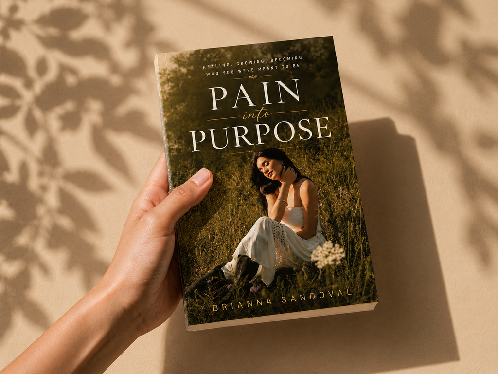A hand holds a paperback book titled 'Pain into Purpose' by Brianna Sandoval. The book cover features a woman sitting in a grassy field, and the book is photographed against a warm beige background with soft leaf shadows.