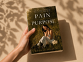 A hand holds a paperback book titled 'Pain into Purpose' by Brianna Sandoval. The book cover features a woman sitting in a grassy field, and the book is photographed against a warm beige background with soft leaf shadows.