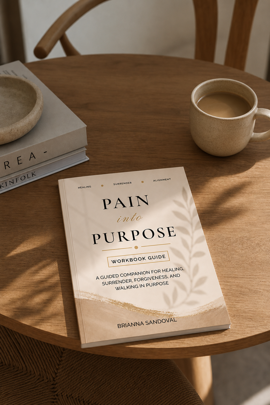 A workbook titled 'PAIN in PURPOSE' by Brianna Sandoval lies on a round wooden table, styled with a cup of coffee, a small bowl, and another book in a warm, sunlit indoor setting.